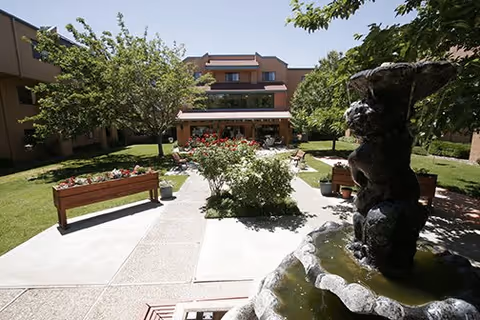Outdoor courtyard area at Sandia Vista Senior Living featuring a stone water fountain in the foreground, paved walkways, benches, green grass, trees, and a building with balconies in the background under a clear sky.