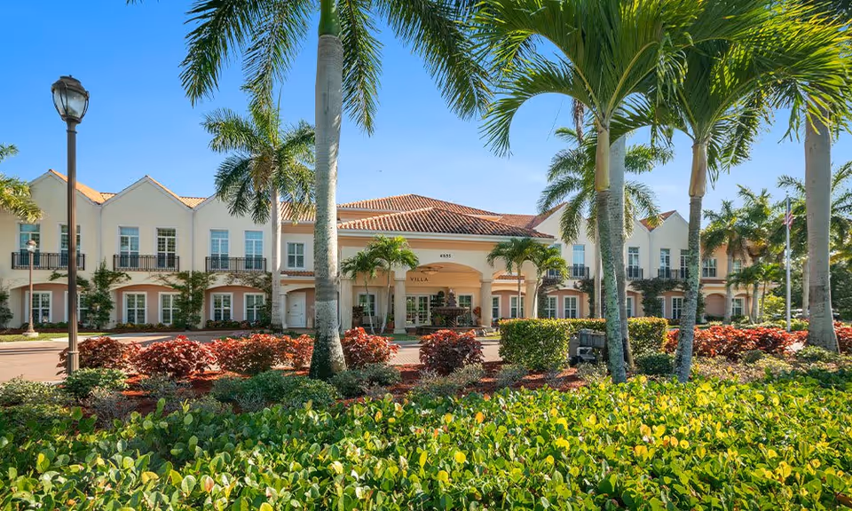 Front exterior view of Villa at Terracina Grand, a two-story building with a tiled roof, surrounded by palm trees and landscaped greenery under a clear blue sky.