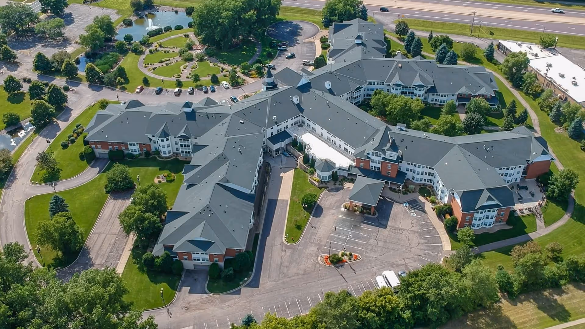 Aerial view of The Rivers Retirement Community showing a large, multi-wing building with dark gray roofs surrounded by green lawns, trees, and landscaped gardens. There is a parking lot in front of the building and a pond with walking paths visible in the upper left corner.