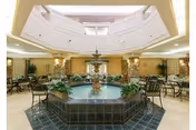 Spacious indoor common area with a large octagonal water fountain in the center surrounded by metal chairs and tables. The room features a high ceiling with skylights and stone pillars, creating a bright and inviting atmosphere.