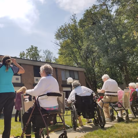 A group of elderly people sitting outdoors on chairs and wheelchairs in a garden area near a two-story building, with trees and a partly cloudy sky in the background. A caregiver or staff member stands nearby wearing a teal and black shirt.