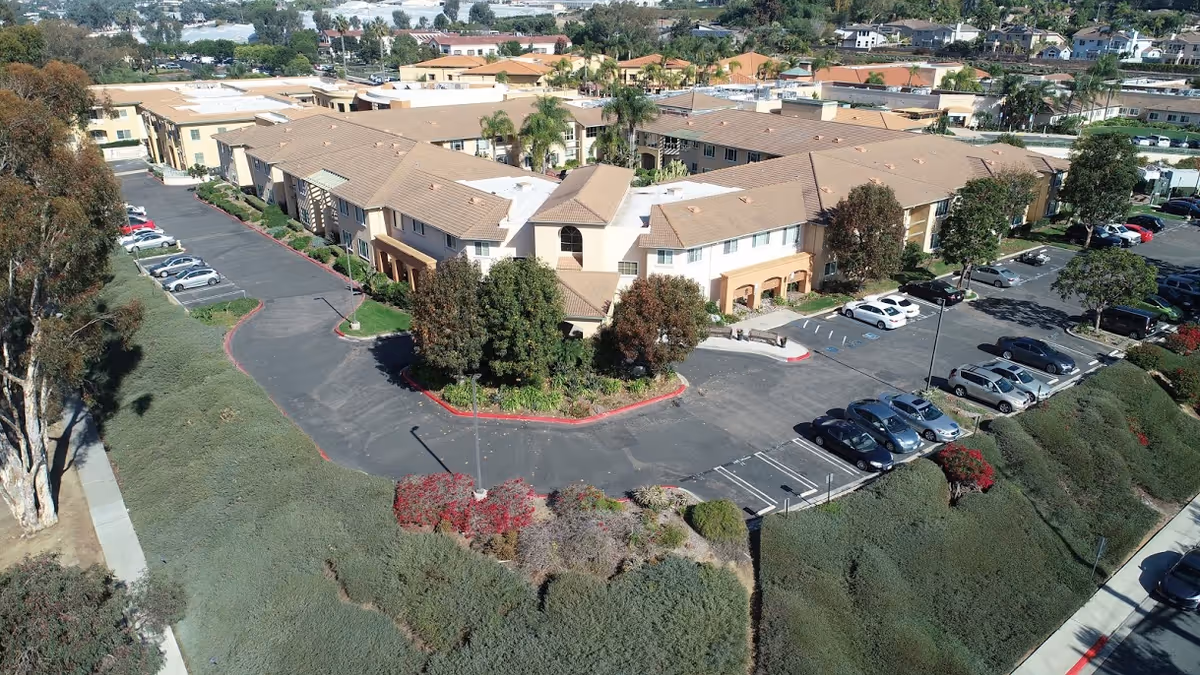 Aerial view of a large retirement community building with beige walls and tan roofs, surrounded by parking lots with several parked cars and landscaped greenery including trees and shrubs.