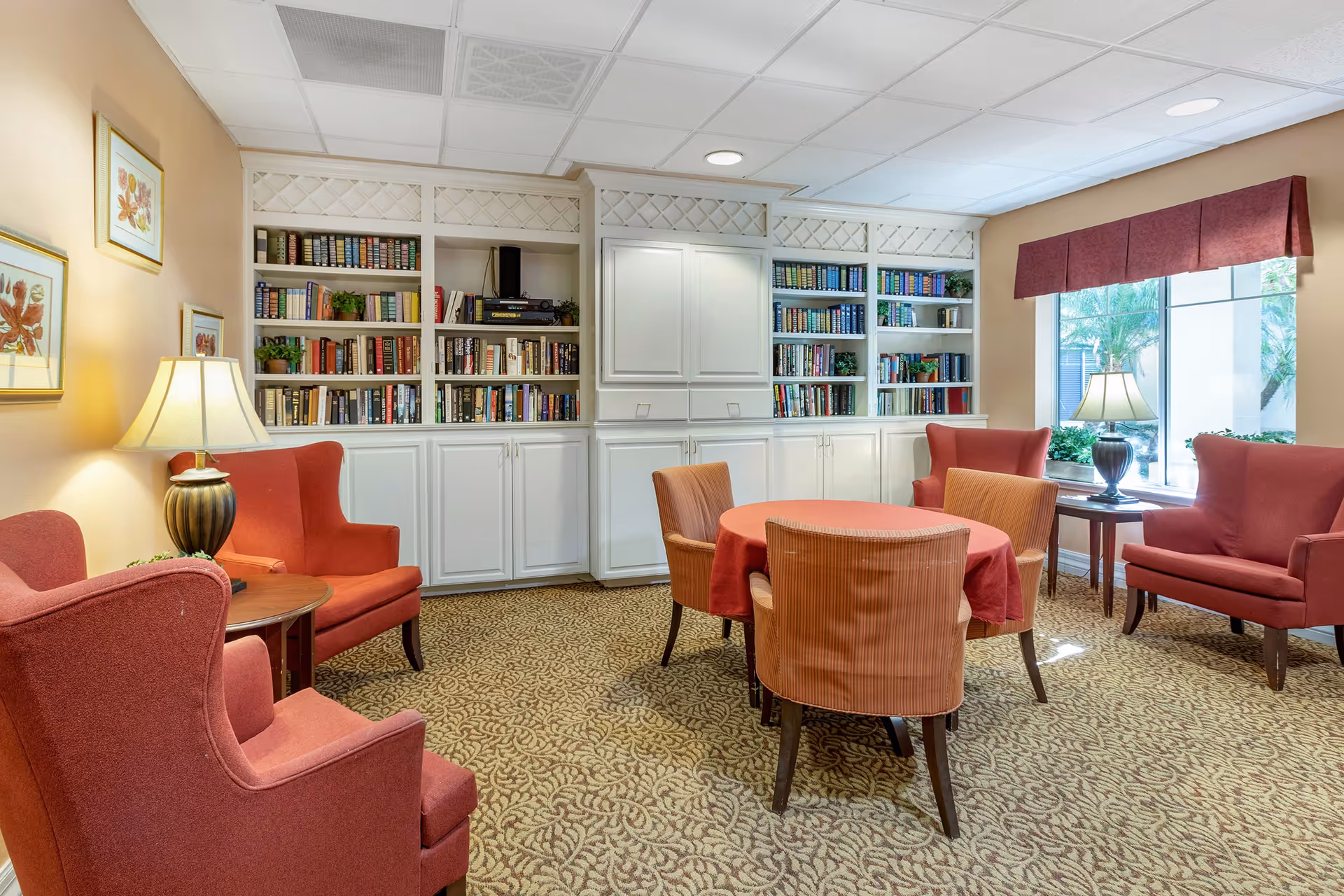 Common sitting room with built-in bookshelves, upholstered chairs around a round table, and lamps by a window.