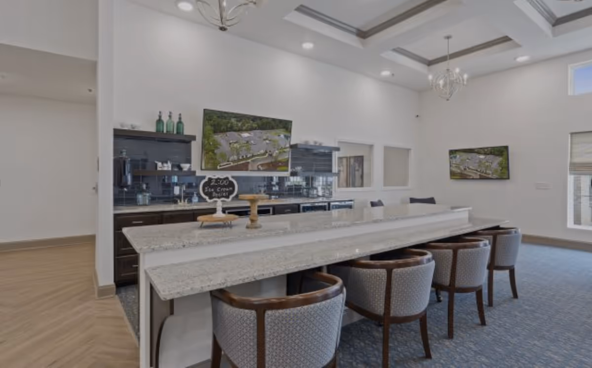 Bright communal dining and bar area with a long marble counter, upholstered barstools, cabinetry and wall-mounted TVs.