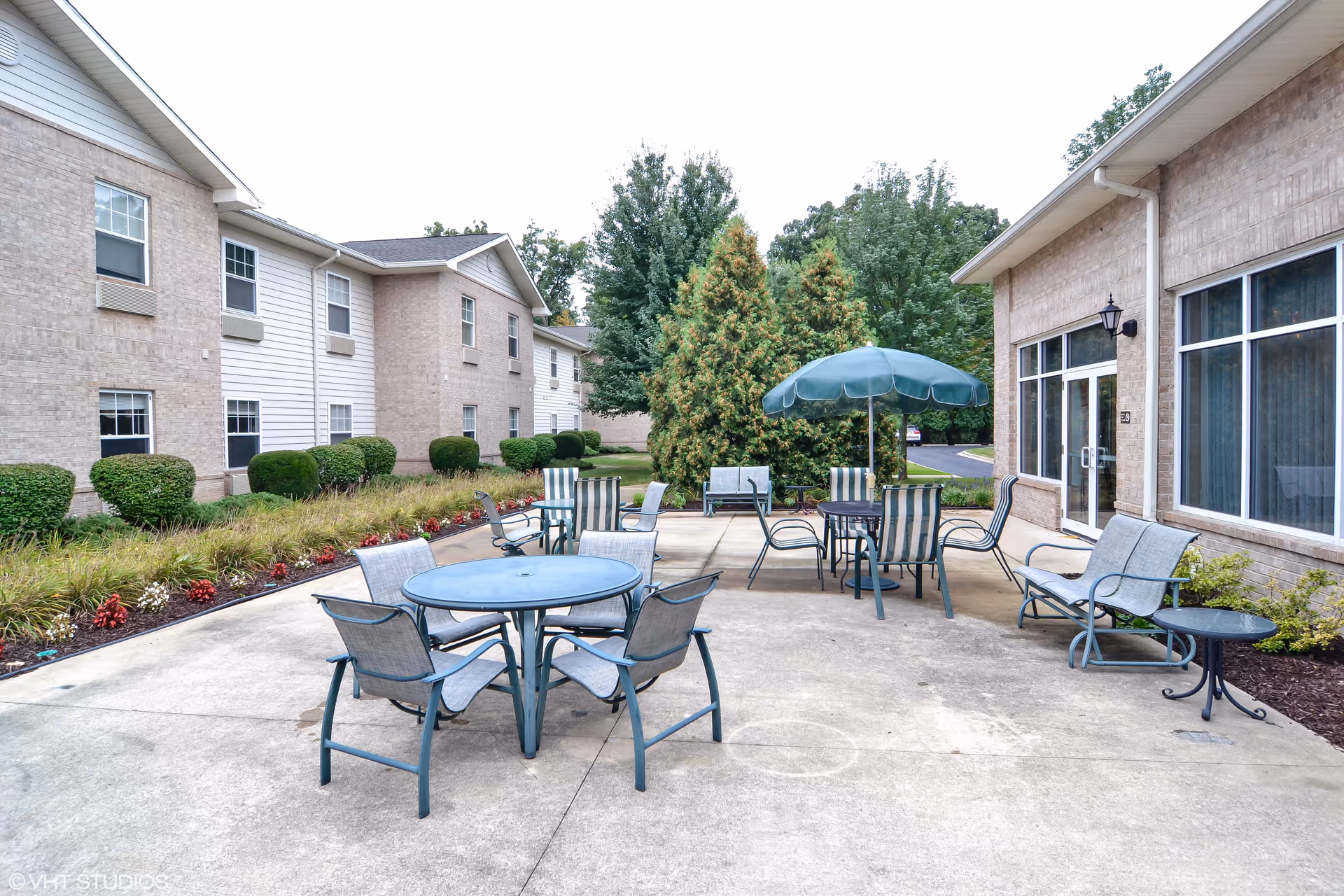 Outdoor patio area at Cedarhurst Senior Living of Edison Lakes with multiple tables and chairs, including one table with a green umbrella. The patio is surrounded by shrubs and trees, with two beige brick buildings visible on either side.