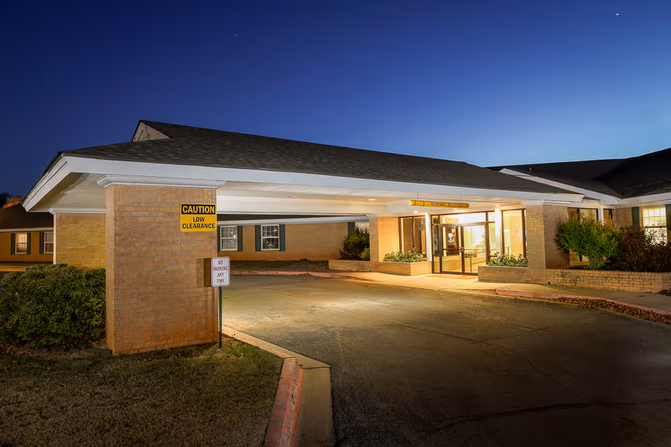 Exterior nighttime view of the entrance to a brick building with a covered driveway and illuminated glass doors. There are caution and no parking signs near the entrance, and some bushes and windows are visible on the building.