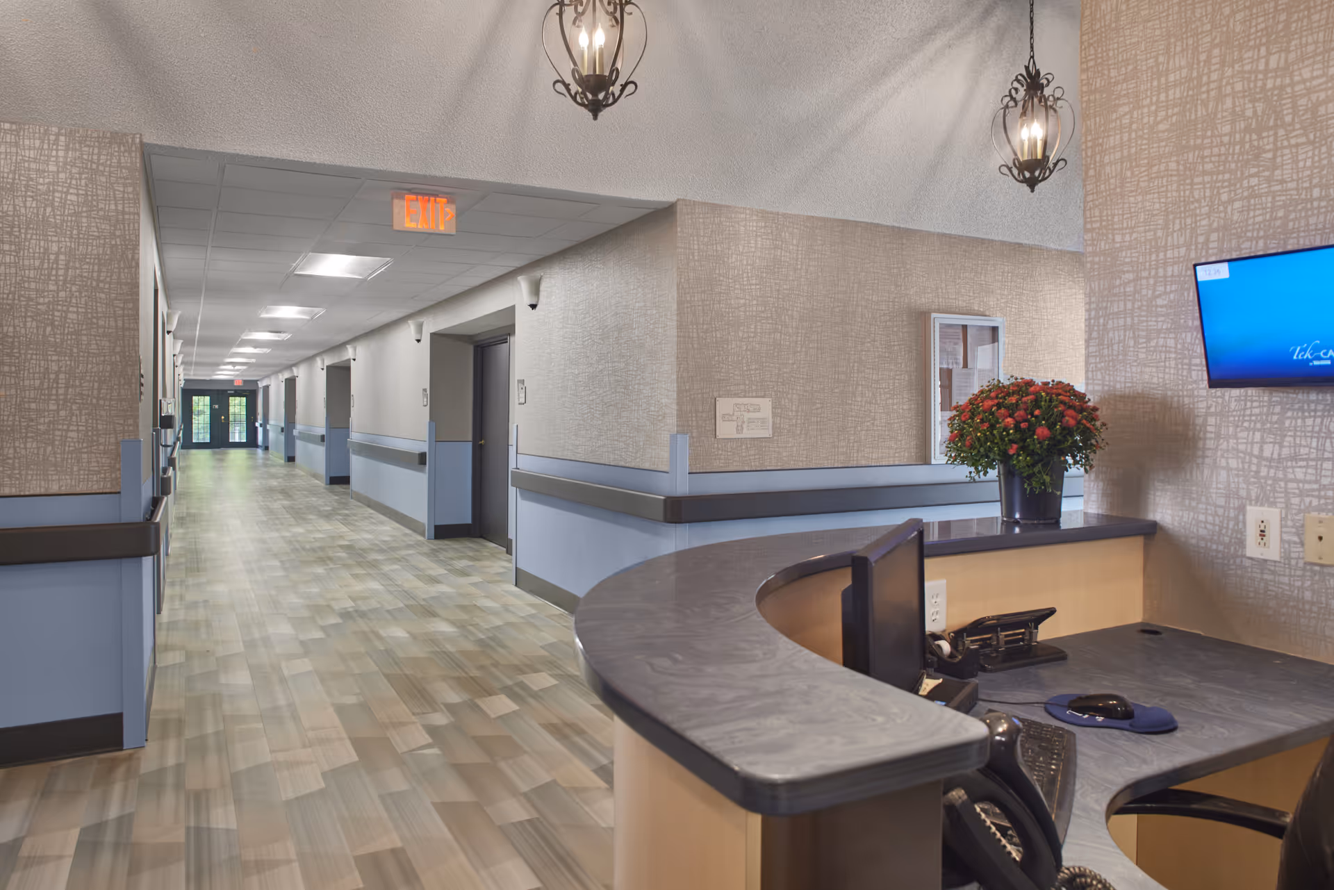 Interior view of a senior living facility hallway with a reception desk in the foreground. The hallway has light-colored walls with a blue accent strip and multiple doors along the corridor. The reception desk has a computer monitor, phone, and a vase with red flowers. Two decorative hanging light fixtures are visible above the desk, and an exit sign is illuminated at the end of the hallway.