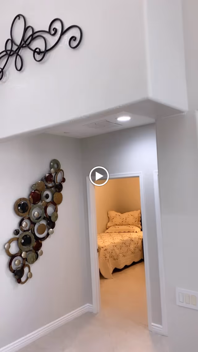 View of a hallway with a decorative wall art piece made of circular elements on the left wall. The hallway leads to a bedroom with a bed covered in a beige quilt and matching pillows. The walls are painted white and the floor is tiled.