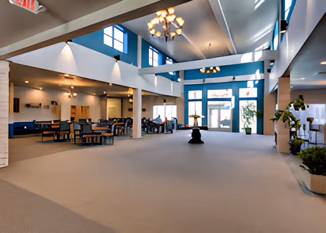 Spacious interior common area of Rock Island Village Assisted Living with high ceilings, large windows, chandeliers, seating areas with tables and chairs, and potted plants near the entrance.