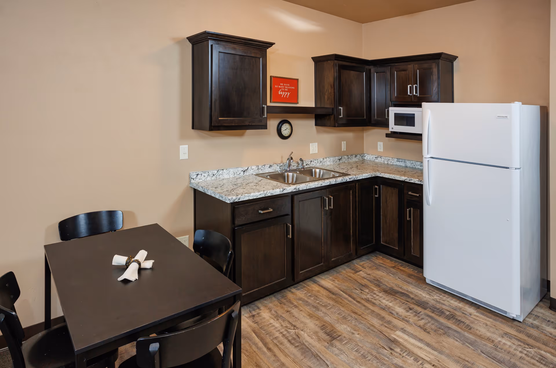 A small kitchen area with dark wood cabinets, a white refrigerator, a microwave, a double sink, and a black dining table with four chairs. The table has two rolled napkins with napkin rings placed on it. The floor is wood-patterned, and the walls are beige with a small red framed sign and a clock above the sink.