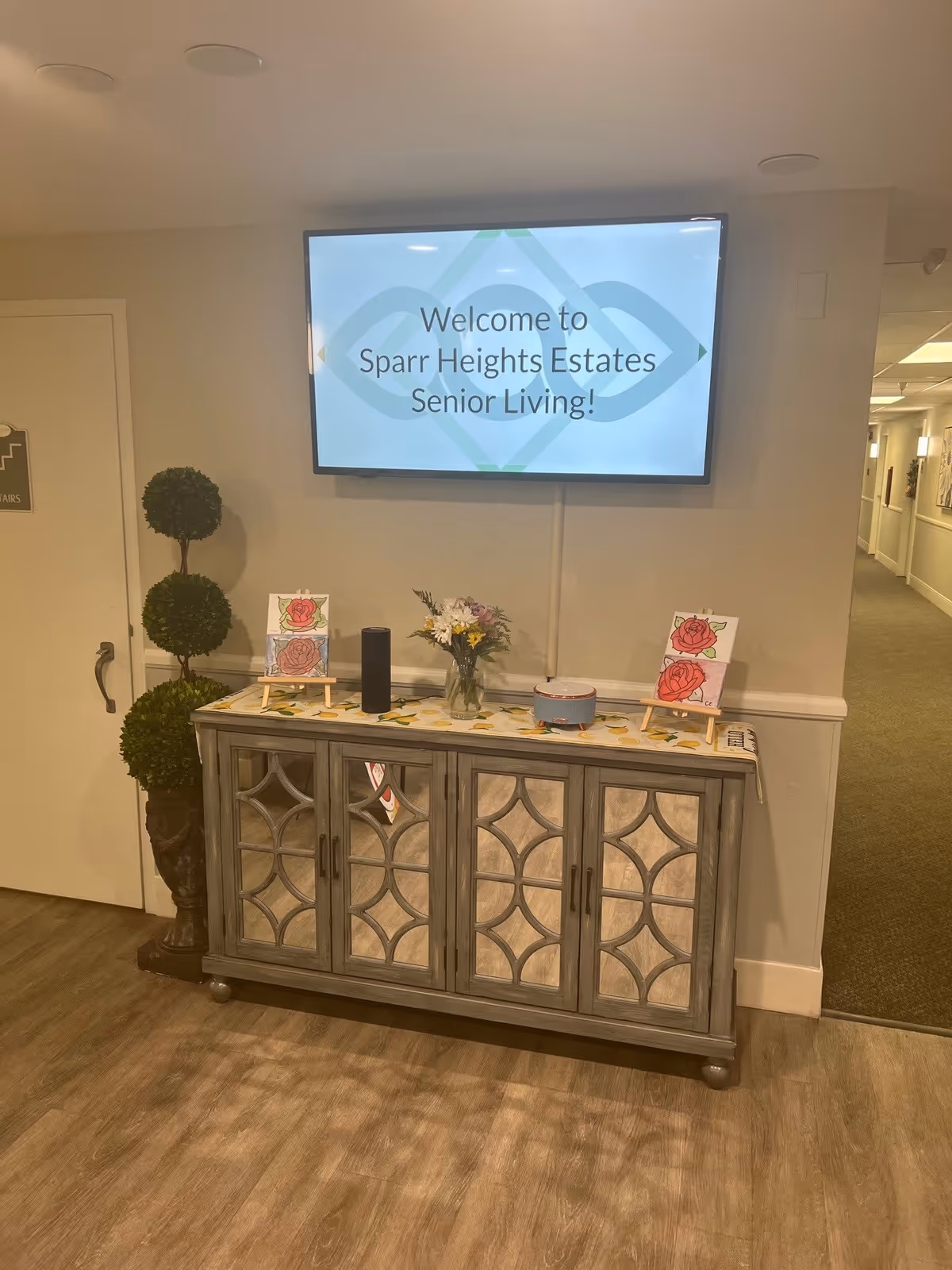 Interior view of a senior living facility hallway with a decorative cabinet against the wall. On top of the cabinet are two small easels holding drawings of roses, a vase with flowers, a black cylindrical speaker, and a small round container. Above the cabinet is a mounted flat-screen TV displaying the message 'Welcome to Sparr Heights Estates Senior Living!'. To the left of the cabinet is a decorative plant with three round green topiaries. The hallway extends to the right with carpeted flooring and wall decorations.