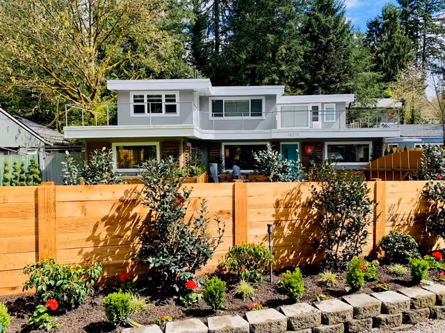 A modern two-story house with large windows surrounded by tall trees. In the foreground, there is a wooden fence with a landscaped garden featuring various shrubs and small plants. The sky is clear and blue.