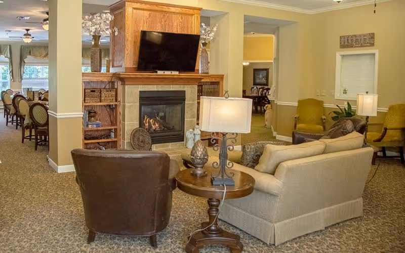 Cozy communal living area with sofas and chairs arranged around a fireplace and TV, with a dining area visible in the background.