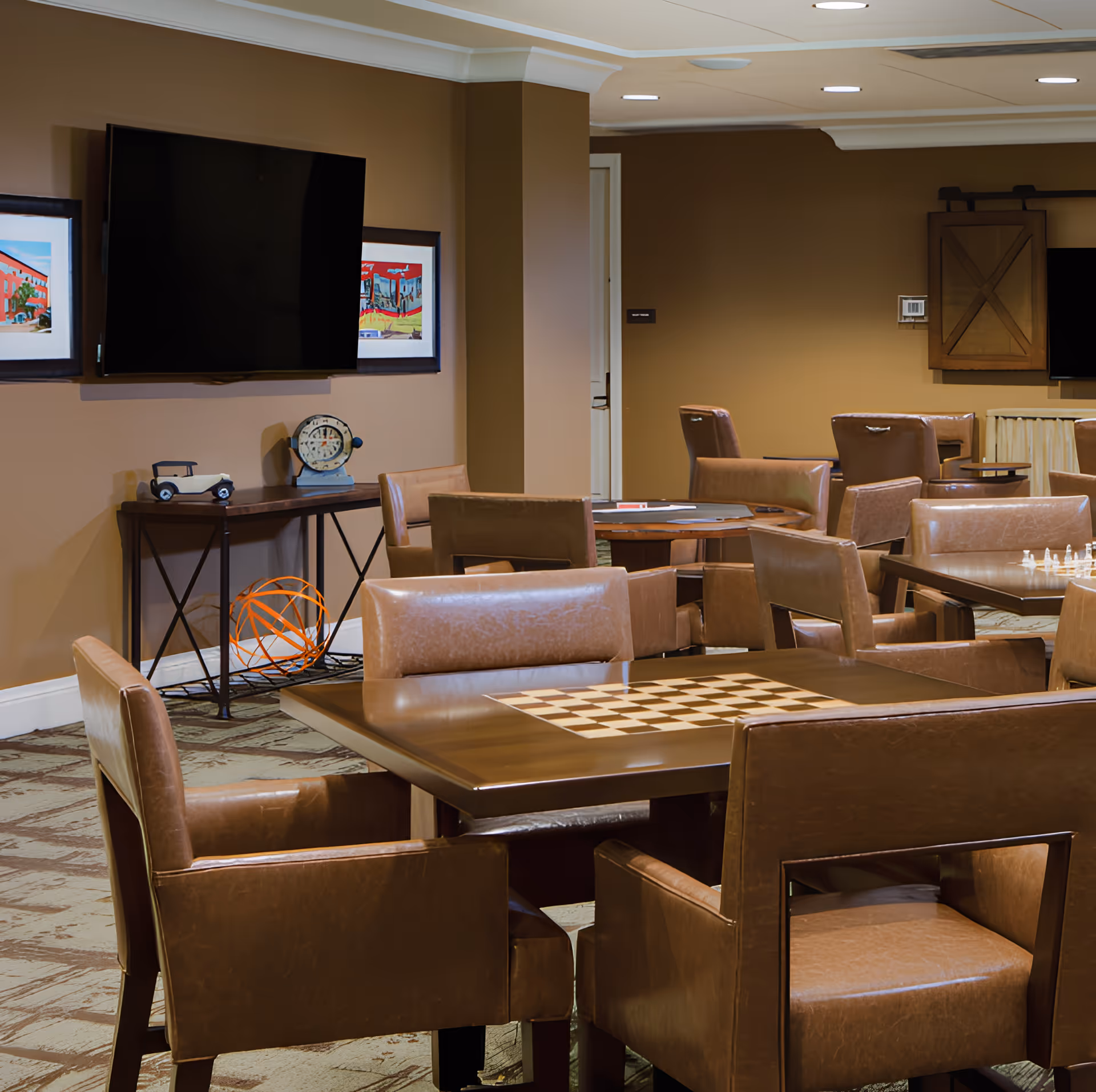 Community activity room with leather chairs and tables (one with a chess/checkerboard), a wall-mounted TV, and decor on the walls.