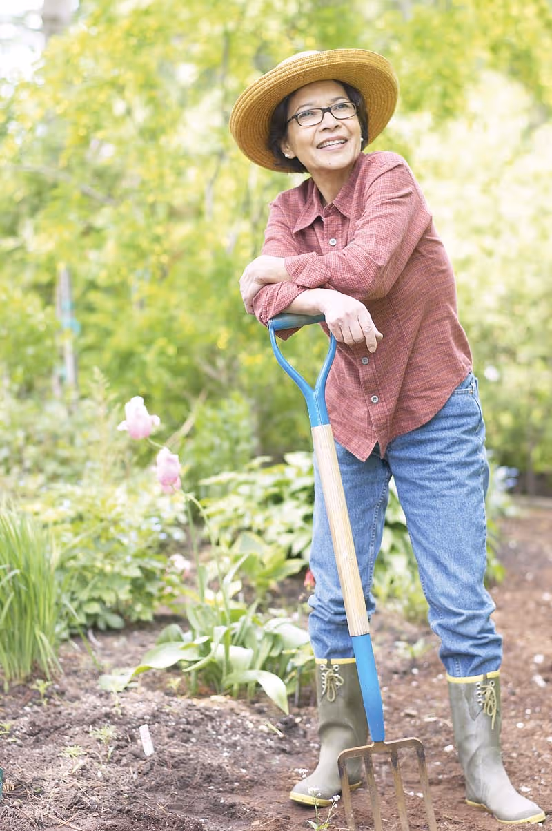 An elderly woman wearing a straw hat, glasses, a red plaid shirt, blue jeans, and green rubber boots is standing in a garden. She is smiling and leaning on a garden fork with plants and flowers visible in the background.