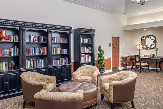 Cozy senior living lounge with bookshelves, four upholstered armchairs around a central ottoman and a desk area.