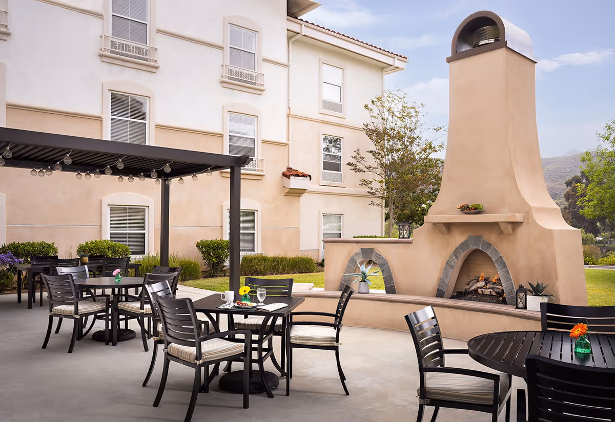 Outdoor patio area at Belmont Village Senior Living Sabre Springs featuring several black tables and chairs with cushions, a large beige outdoor fireplace with a fire burning, string lights hanging from a pergola, and a beige building with multiple windows in the background.