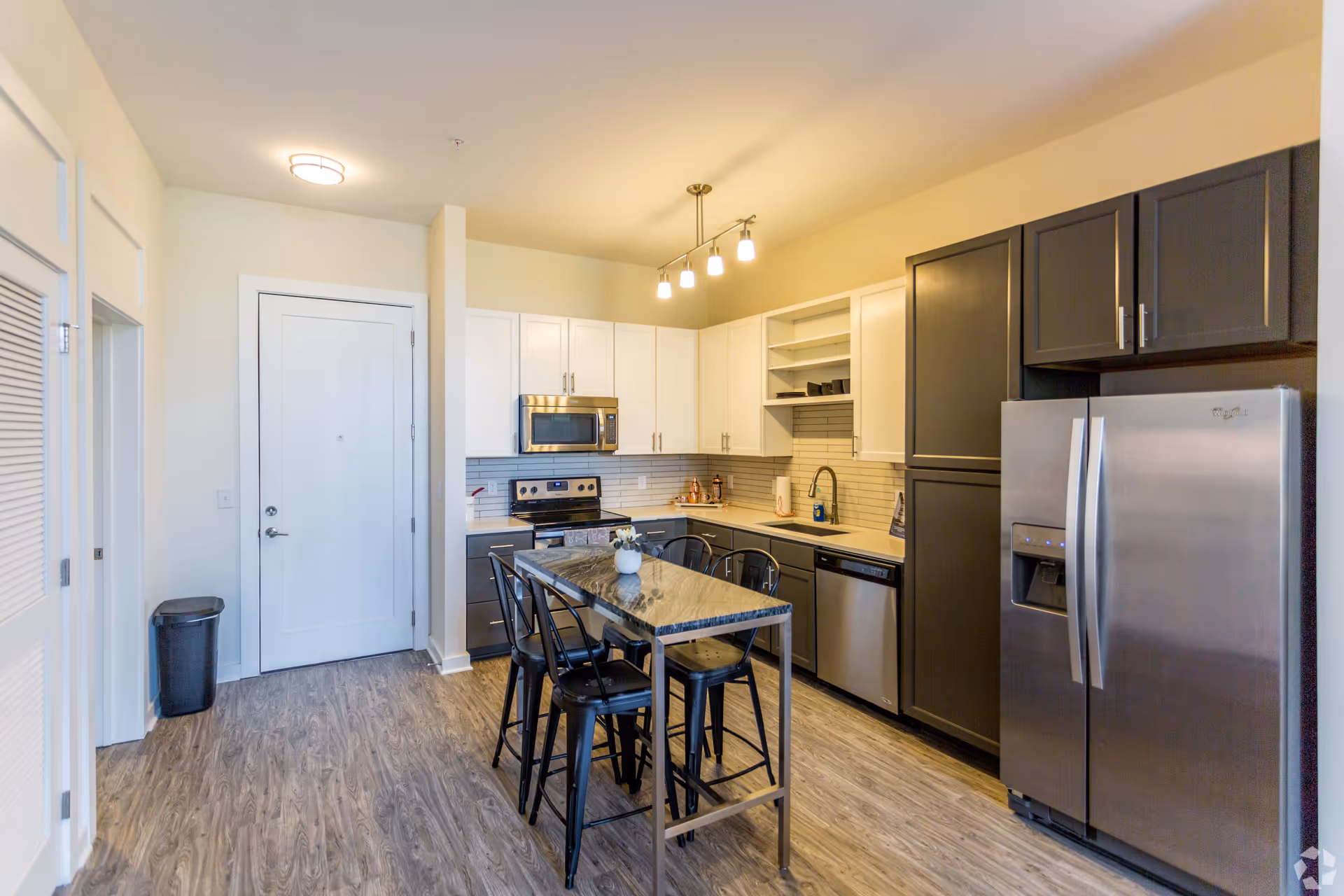 Modern kitchen with a mix of white and dark gray cabinets, stainless steel appliances including a refrigerator, microwave, stove, and dishwasher. A rectangular kitchen island with a marble-like countertop and four black metal chairs is centered in the room. The floor has wood-like vinyl planks, and the walls are painted a light beige color. A ceiling light fixture with four bulbs hangs above the island.