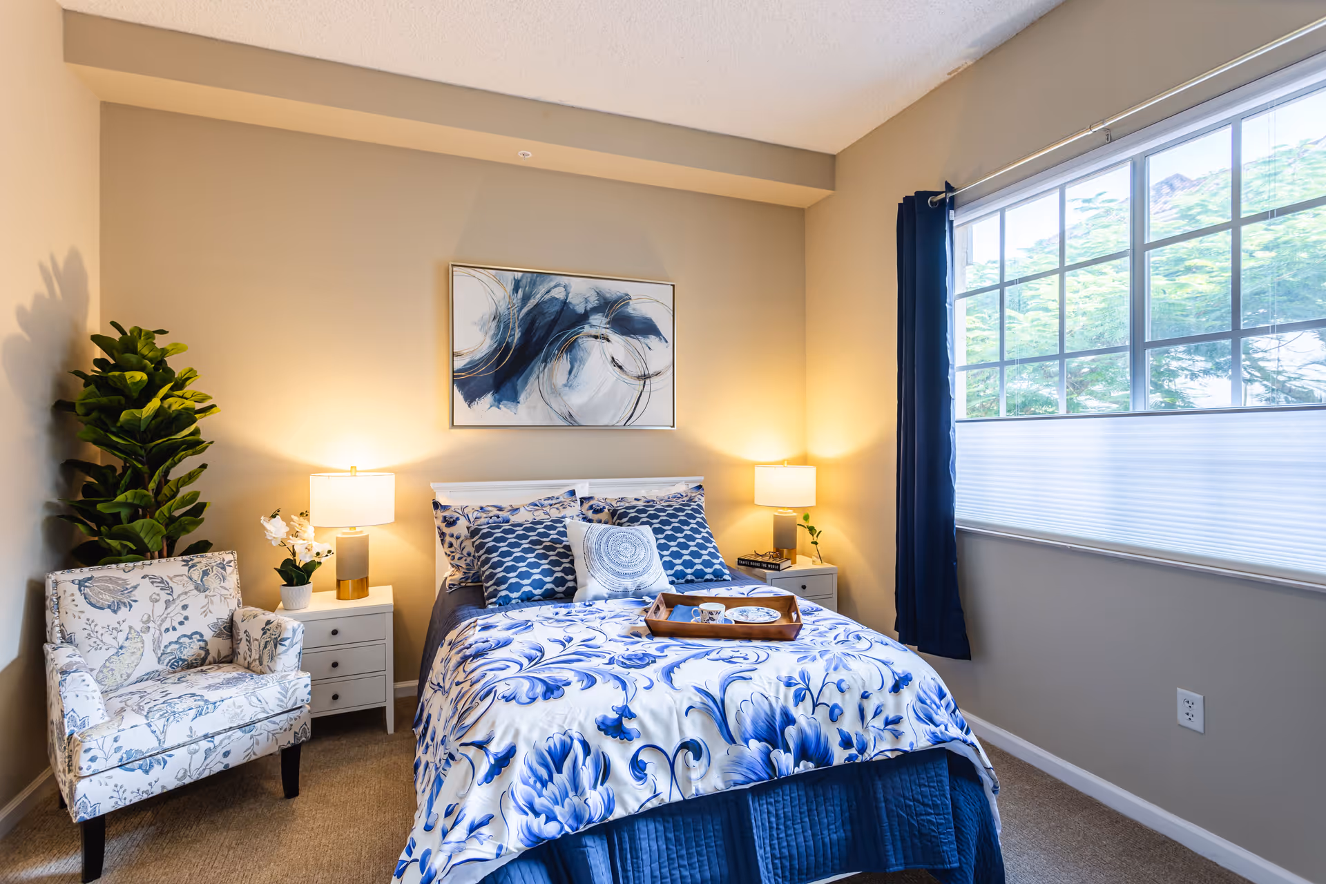 A cozy bedroom with a bed featuring blue and white floral bedding and multiple pillows. There are two bedside tables with lamps on each side of the bed. A floral patterned armchair and a tall green plant are positioned to the left. A large window with blue curtains is on the right wall, allowing natural light into the room. Above the bed hangs an abstract blue and white painting.