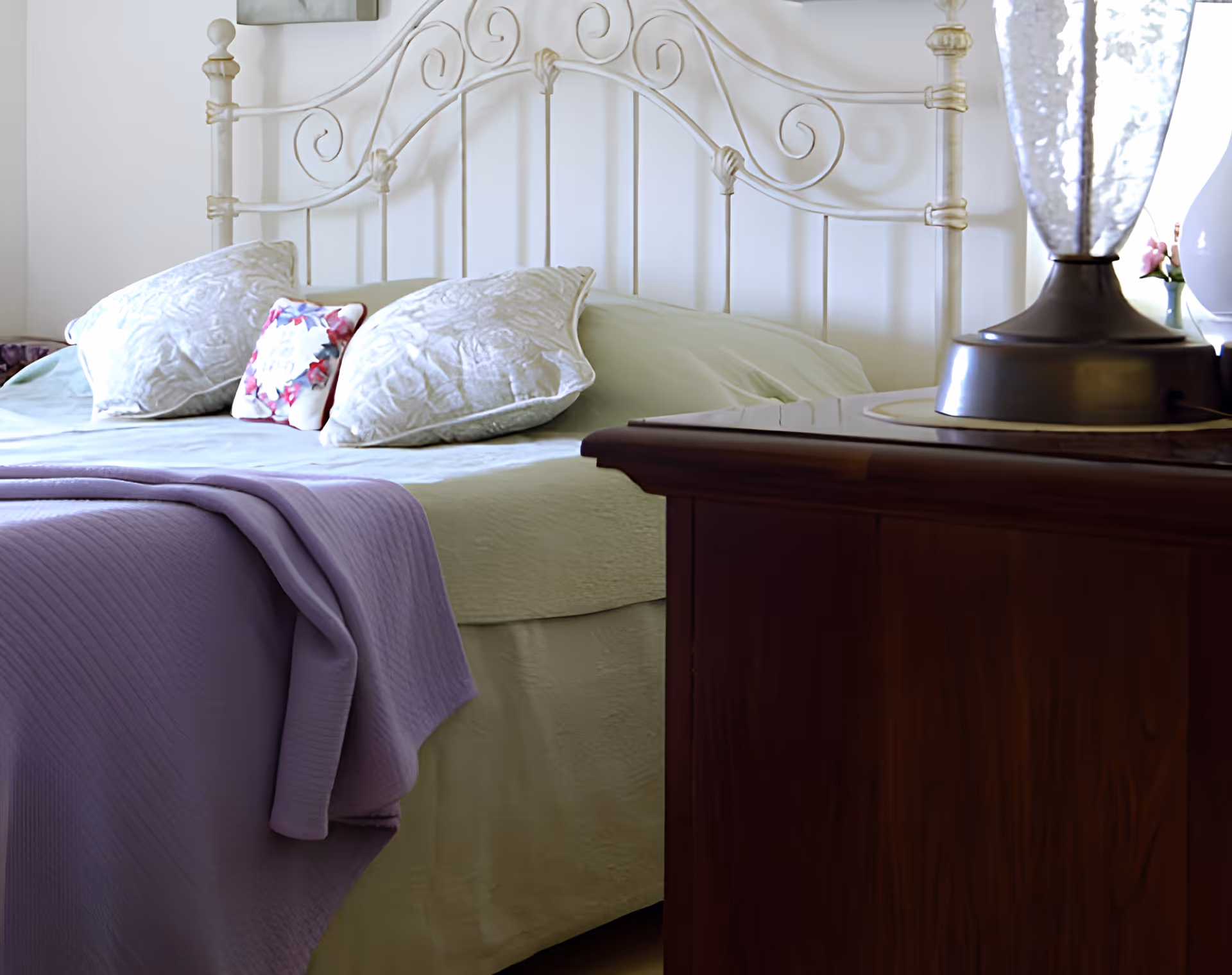 A neatly made bed with a decorative metal headboard, pillows and a purple throw next to a wooden nightstand with a lamp.