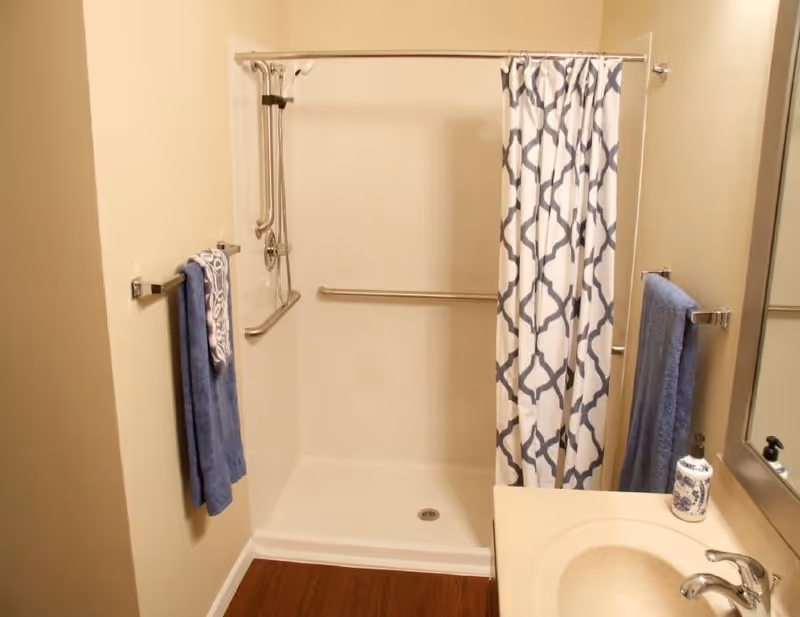 A bathroom shower area with a white shower base and walls, equipped with a silver grab bar and a detachable showerhead. A white and blue patterned shower curtain is partially drawn. Blue towels hang on silver towel racks on both sides of the shower. A beige countertop with a sink and a soap dispenser is visible next to a large mirror.