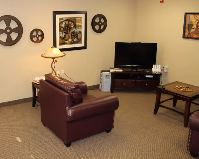 A cozy living room area with a dark brown leather armchair facing a flat-screen TV on a wooden TV stand. The walls are decorated with framed pictures and round wooden wall art resembling film reels. There is a side table with a lamp and a coffee table with a decorative item on it. The carpet is beige and the room has a warm, inviting atmosphere.