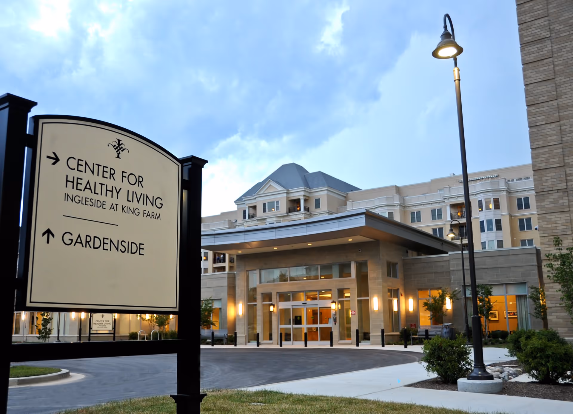 Exterior view of the Ingleside at King Farm building at dusk with a sign in the foreground pointing to the Center for Healthy Living and Gardenside. The building is well-lit with large windows and a covered entrance, surrounded by landscaped greenery and street lamps.