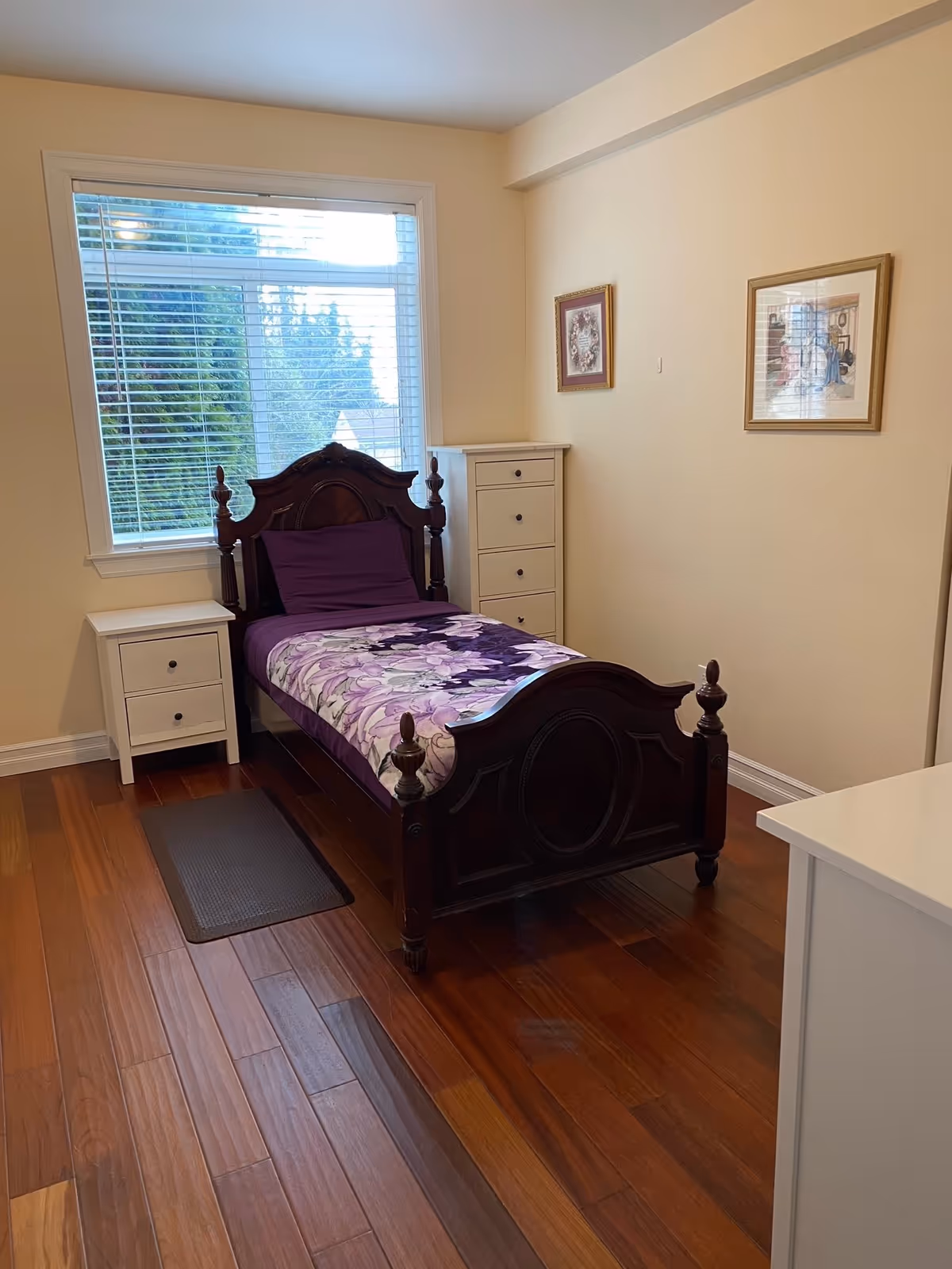 A small bedroom with a single dark wooden bed featuring a purple floral blanket and matching pillow. The room has wooden flooring, a white nightstand with two drawers on the left side of the bed, a tall white chest of drawers in the corner, and two framed pictures hanging on the beige wall. A large window with white blinds is behind the bed, letting in natural light.