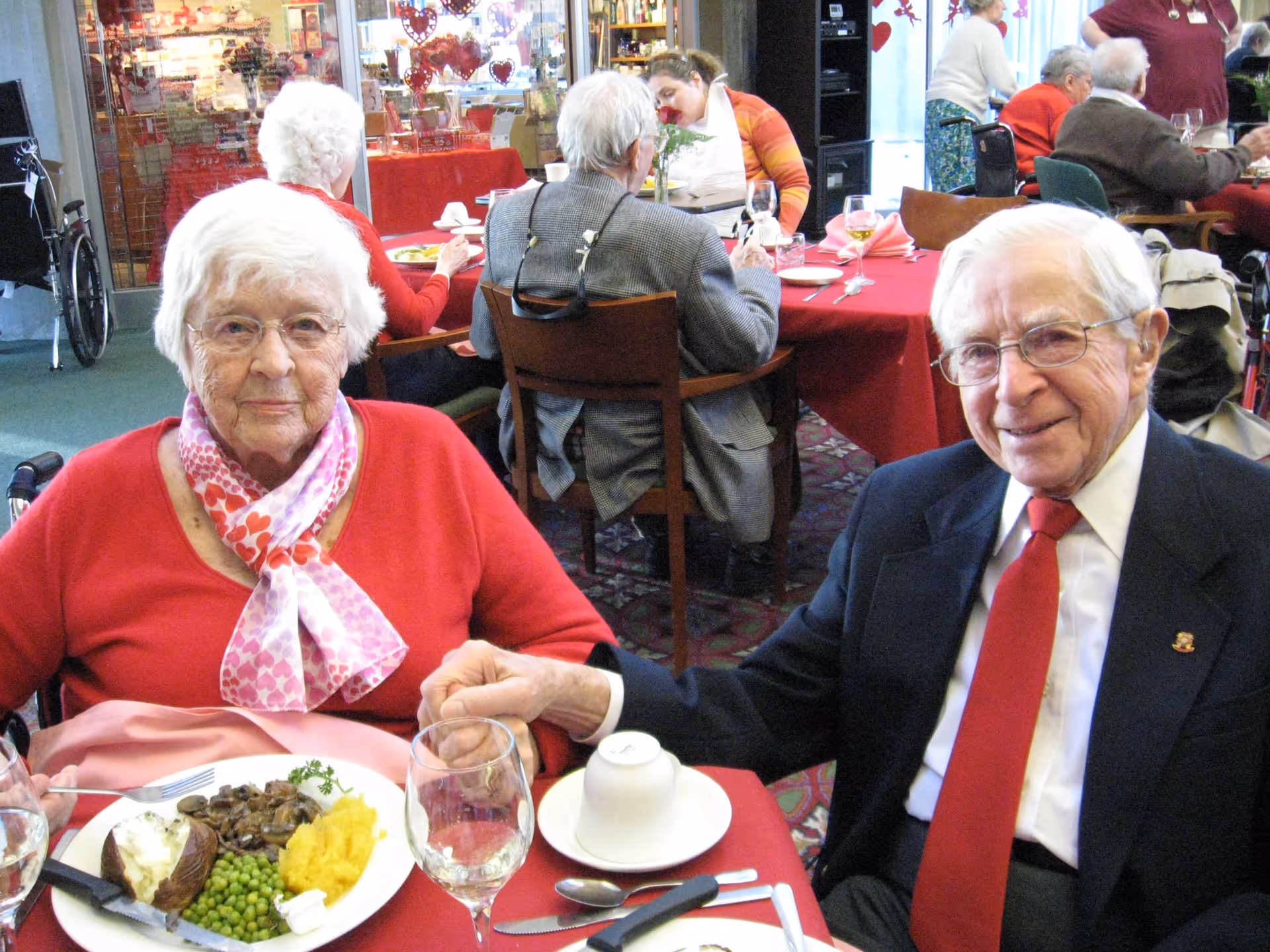 Two elderly residents holding hands and smiling while seated at a red-tableclothed dining table with plates of food in a communal dining room.