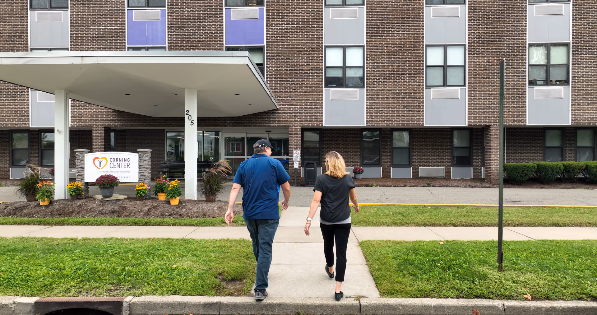 Two people walking on a sidewalk towards the entrance of a multi-story brick building with a covered entrance. A sign near the entrance reads 'Corning Center for Rehabilitation and Nursing'. There are potted plants and greenery around the entrance area.
