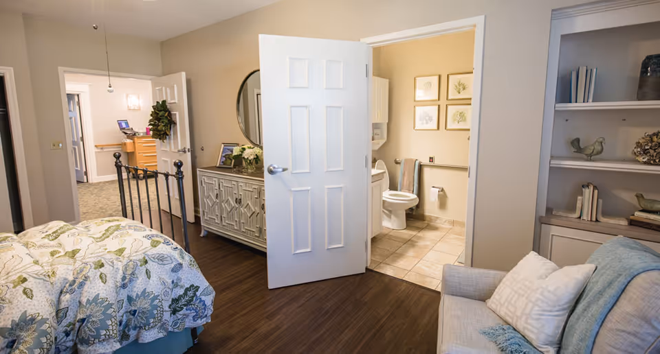 Cozy bedroom with a floral bedspread, dresser and seating area, with an open door showing an en-suite bathroom.