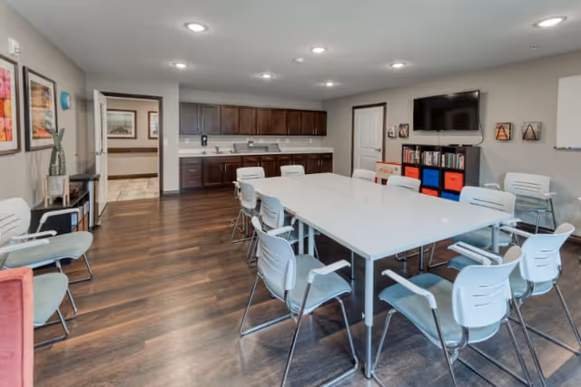 A spacious, well-lit room with a large white rectangular table surrounded by white chairs with light blue cushions. The room has wooden flooring, a kitchenette with dark wood cabinets and a sink along the back wall, and a flat-screen TV mounted on the wall above a bookshelf with colorful storage bins. The walls are decorated with framed pictures and a whiteboard is visible on the right side.