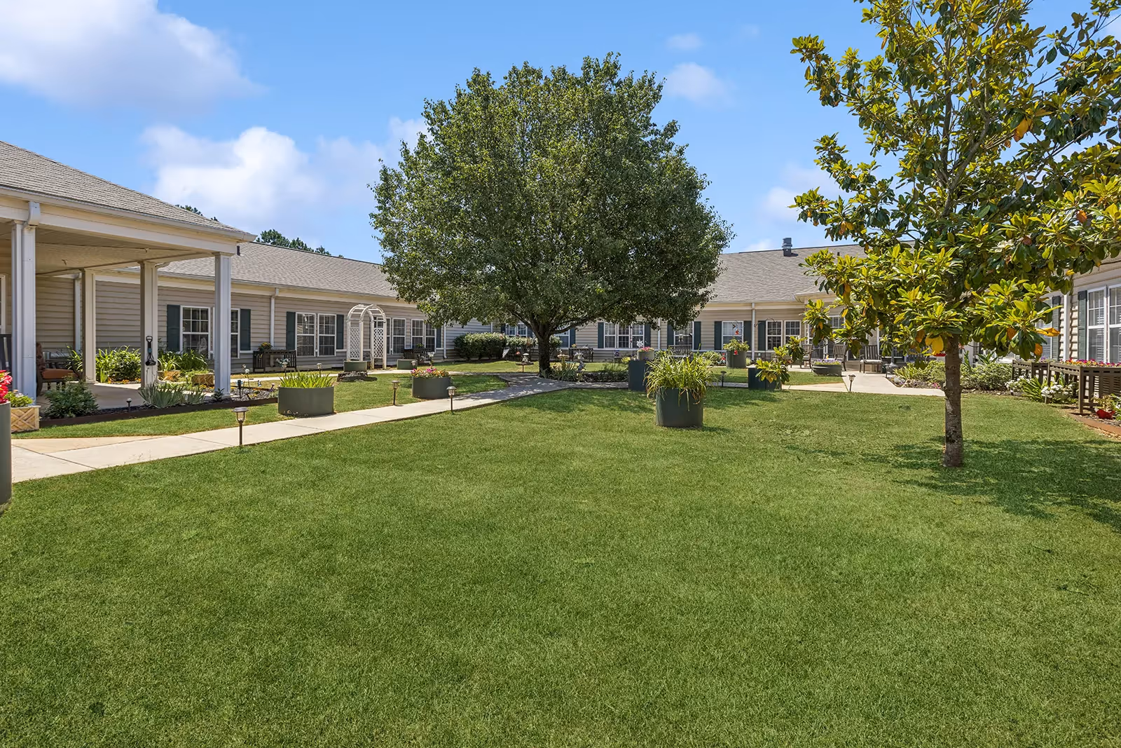 A spacious outdoor courtyard area with green grass, several trees, and large planters. The courtyard is surrounded by a single-story building with beige siding and white trim. There are paved walkways and some outdoor seating areas under covered porches. The sky is clear with a few clouds.