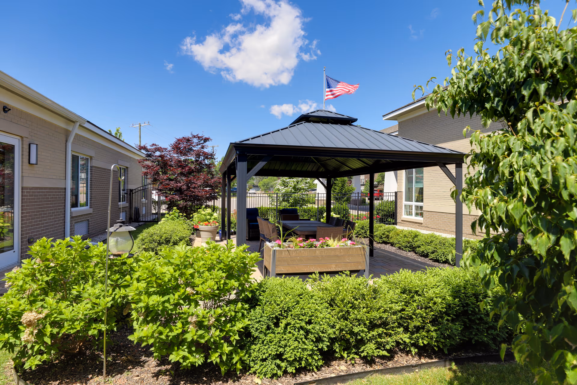 Outdoor garden area at a senior living facility with a black metal gazebo, surrounded by green bushes and plants. There are chairs and a table under the gazebo, and an American flag flying on top. The sky is blue with some clouds, and buildings are visible on either side of the garden.