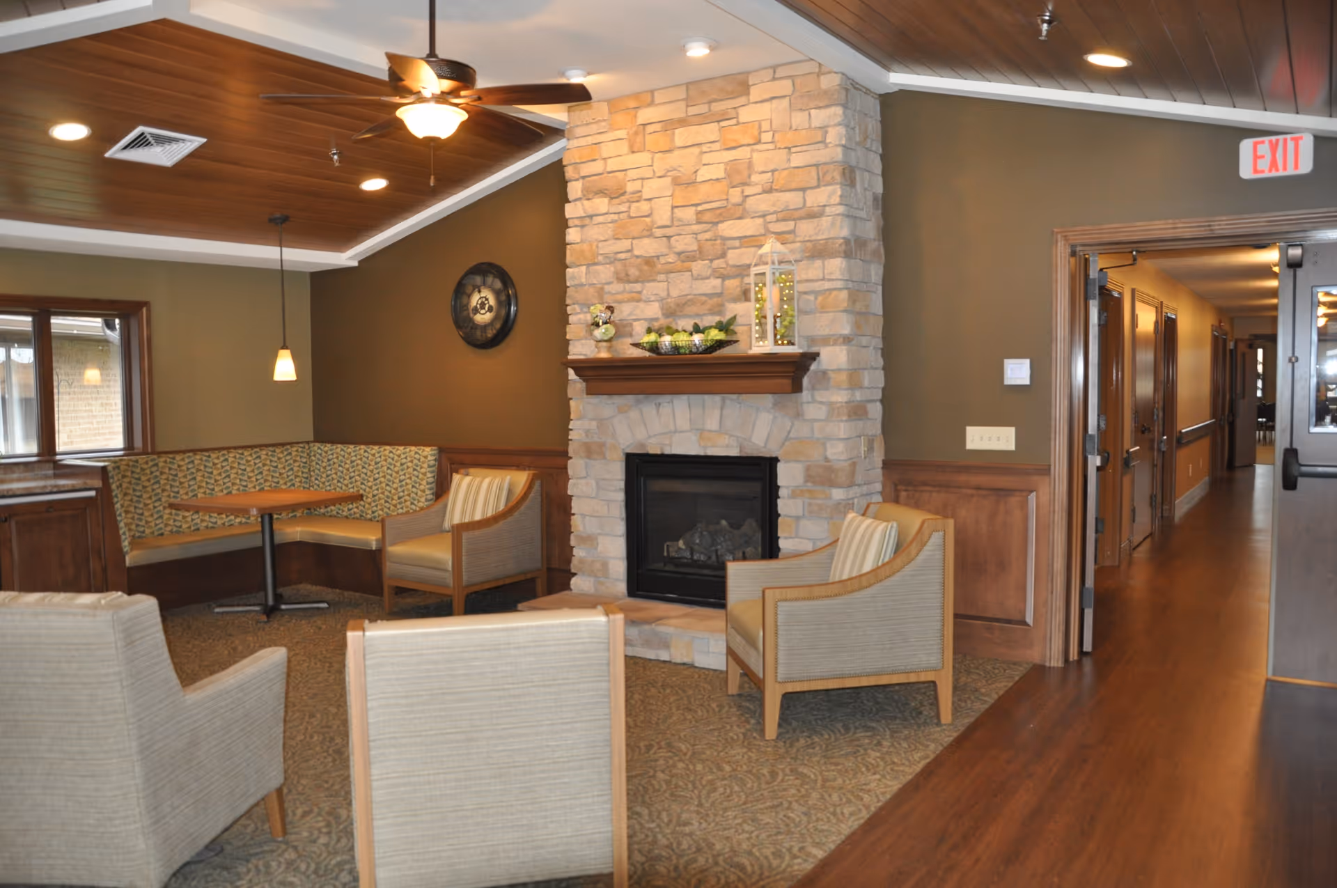 A cozy common area with a stone fireplace, surrounded by four beige armchairs. To the left, there is a built-in corner bench with patterned upholstery and a small wooden table. The ceiling is wooden with recessed lighting and a ceiling fan. A hallway with wooden flooring and doors is visible to the right.