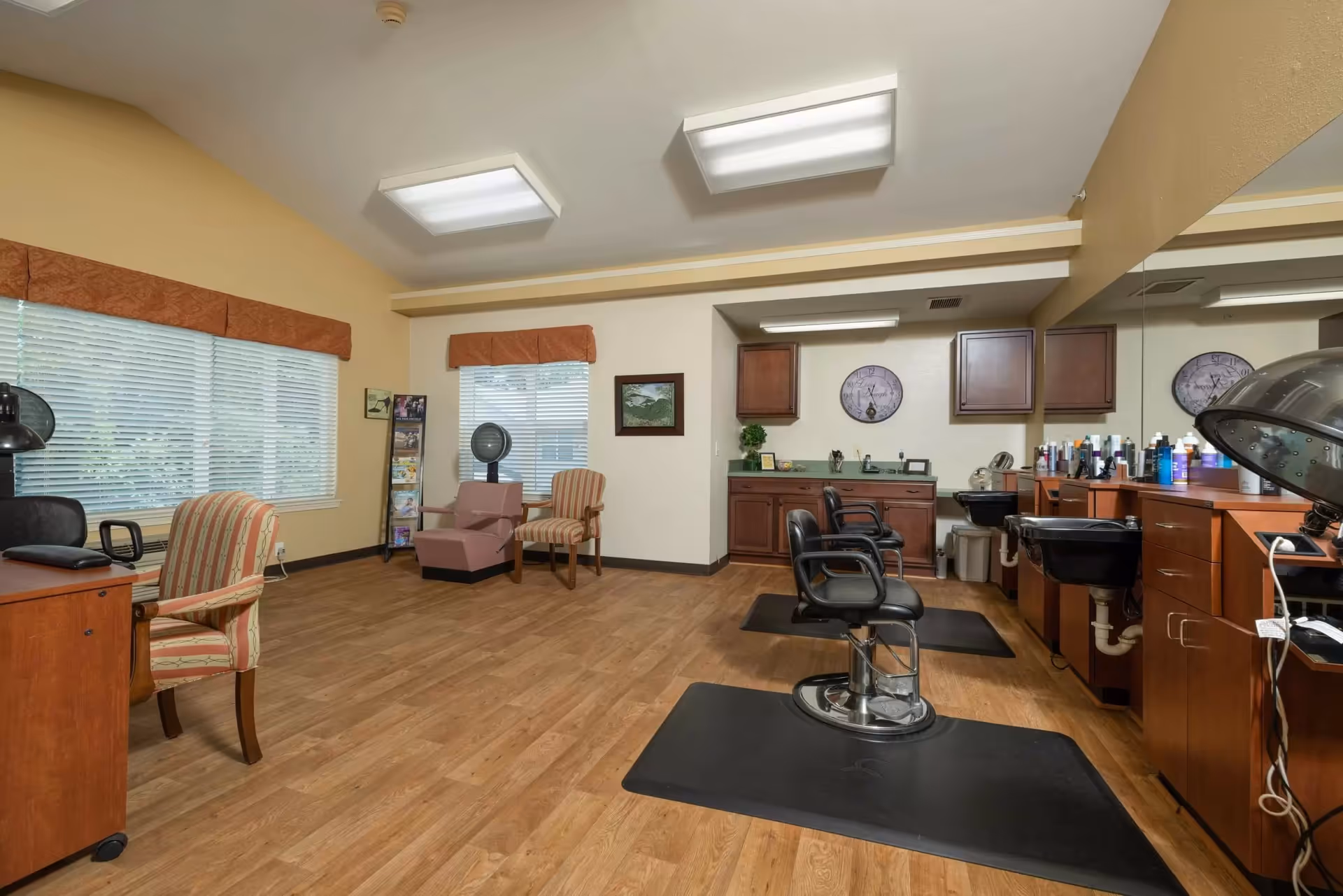 Interior view of a salon area in a senior living facility with wooden flooring, salon chairs, hair washing sinks, and hair drying equipment. The room has large windows with blinds and valances, wooden cabinets, and a few upholstered chairs along the walls.