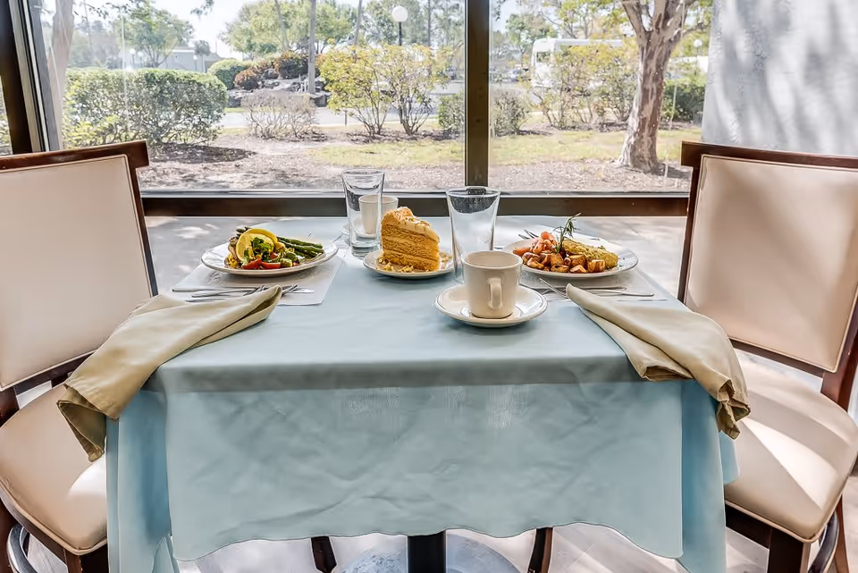 A dining table set for two with a light blue tablecloth and beige napkins on each side. On the table are two plates of food, a slice of cake on a small plate, two glasses of water, and a white coffee cup on a saucer. The table is positioned in front of a large window showing a view of bushes and trees outside.
