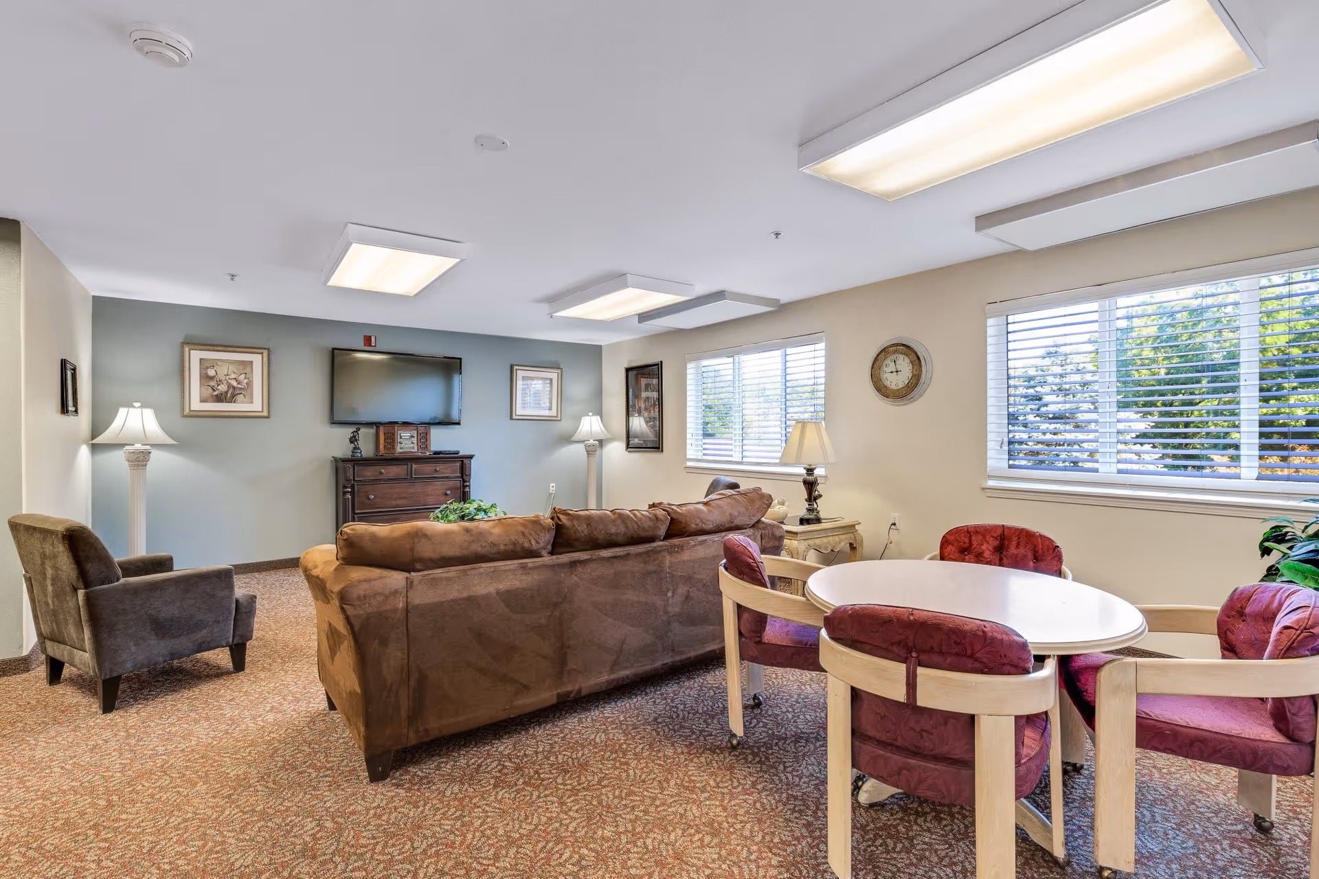 A cozy living room area in a senior living facility with a brown sofa, a dark armchair, a round table with four purple cushioned chairs, two floor lamps, a wall-mounted TV above a wooden cabinet, framed artwork on the walls, and large windows with blinds letting in natural light.