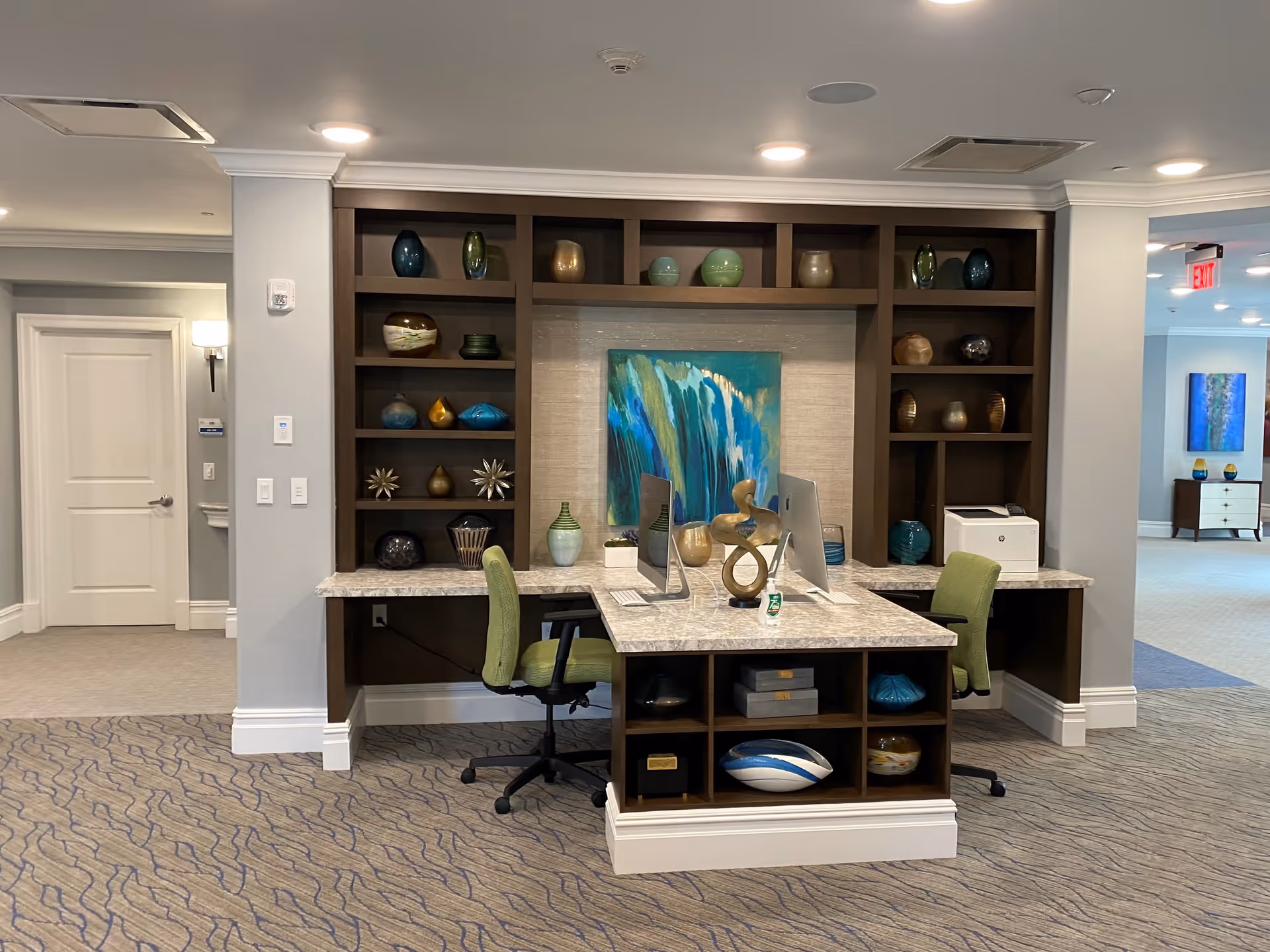 A modern office area within a senior living facility featuring a built-in dark wood shelving unit filled with decorative vases and sculptures. Two green office chairs are positioned at a marble countertop desk with two computer monitors and a small sculpture. The room has beige walls, carpeted flooring with a wavy pattern, and recessed ceiling lights. In the background, there is a hallway with white doors and additional artwork on the walls.