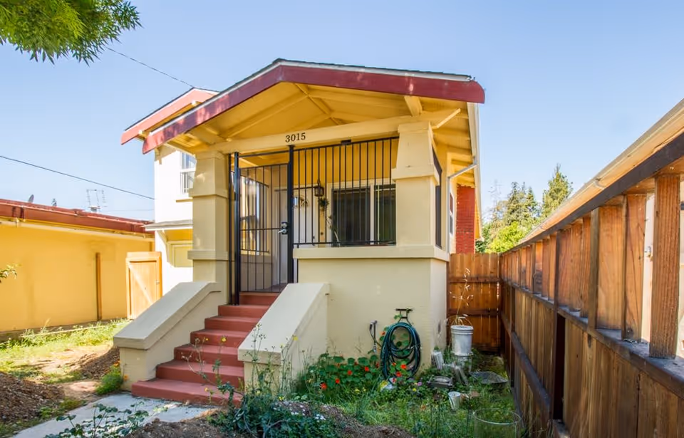 Front exterior view of a small yellow house with a red roof trim and red steps leading up to a gated front porch. There is a garden hose coiled on the right side near the steps and a wooden fence running along the right side of the property under a clear blue sky.