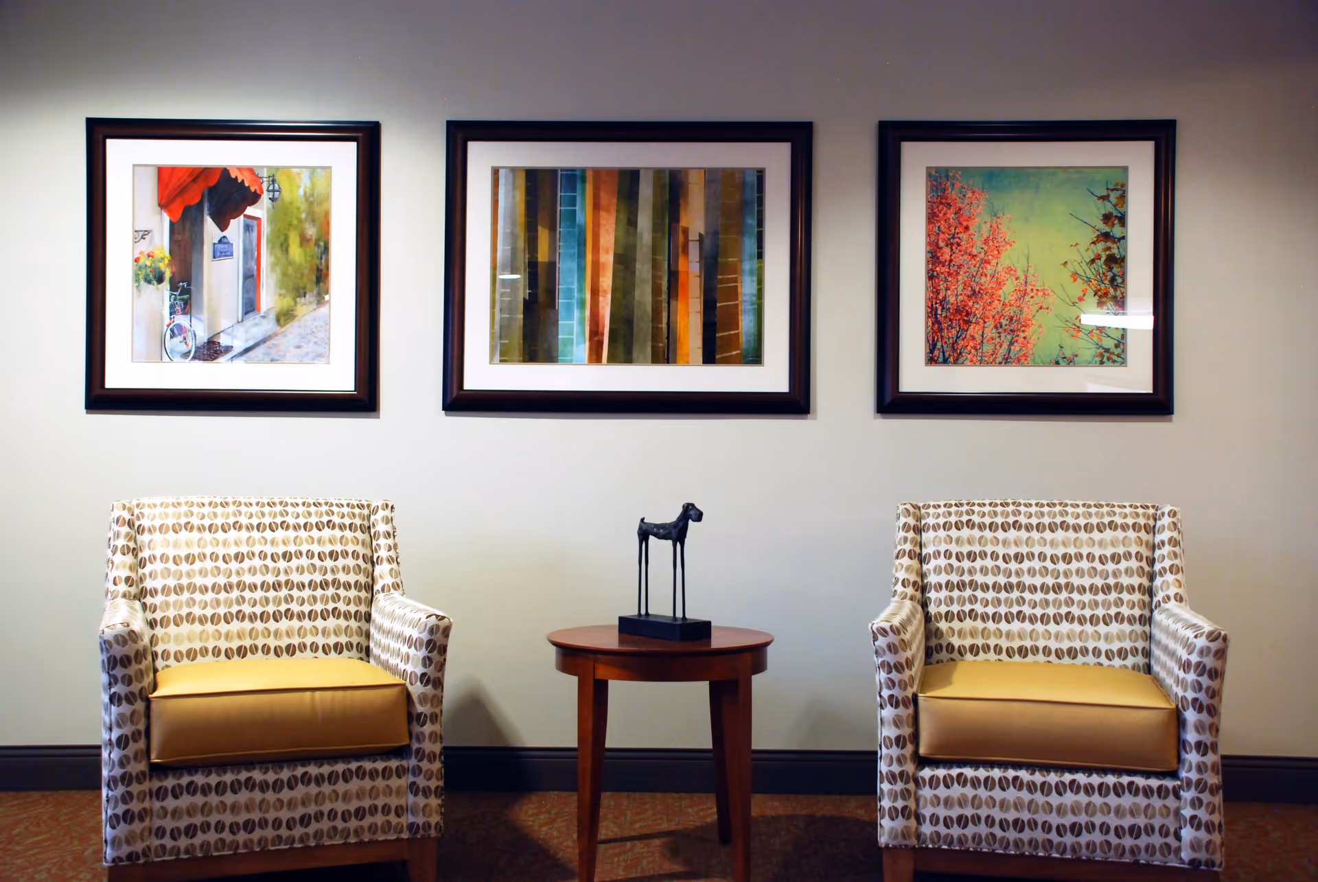 Two patterned armchairs flank a small round table with a decorative sculpture beneath three framed artworks on a wall.
