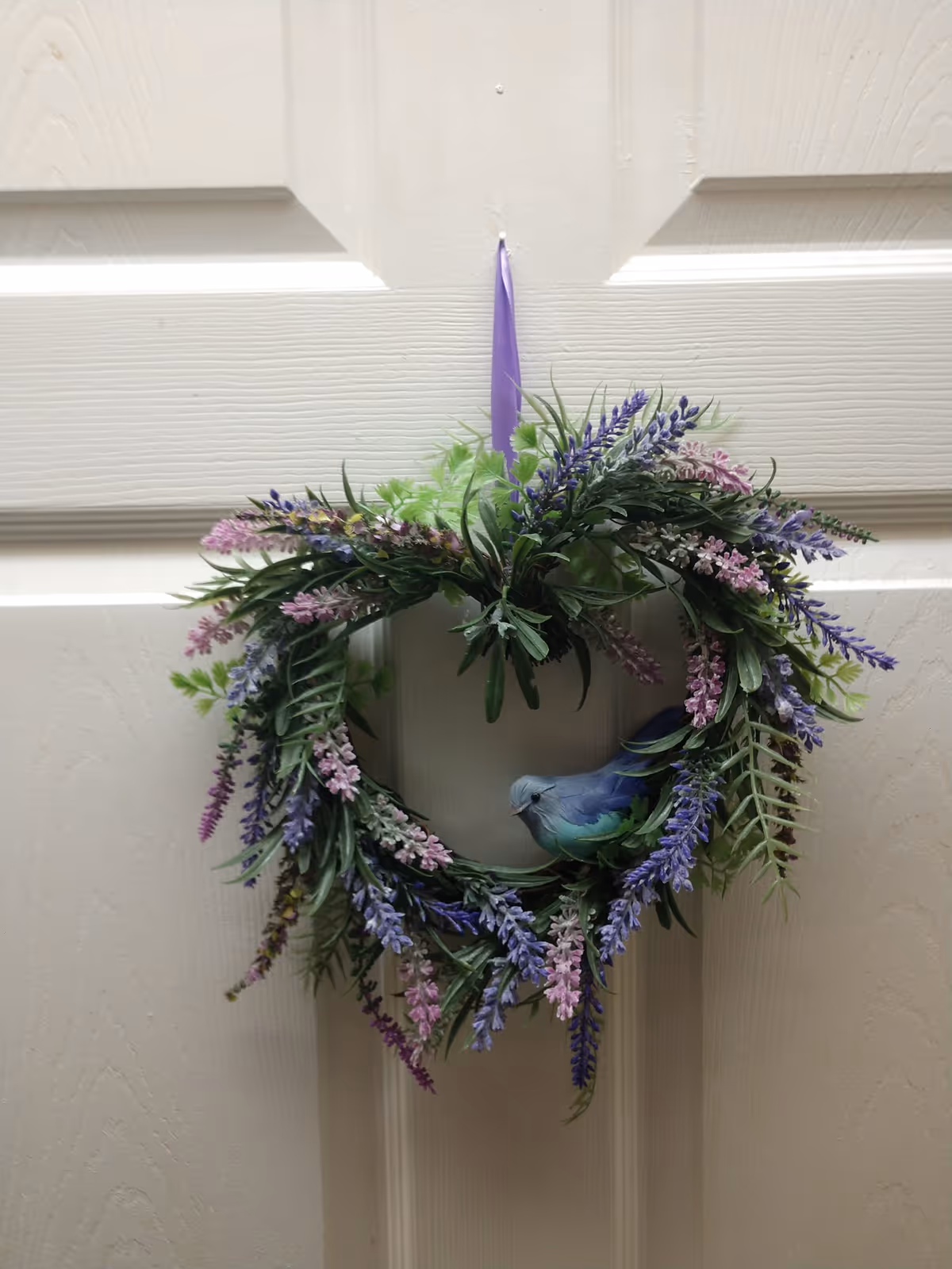 A decorative wreath made of green leaves and purple and pink flowers, with a small blue bird figurine nestled among the foliage, hanging on a white paneled door by a purple ribbon.