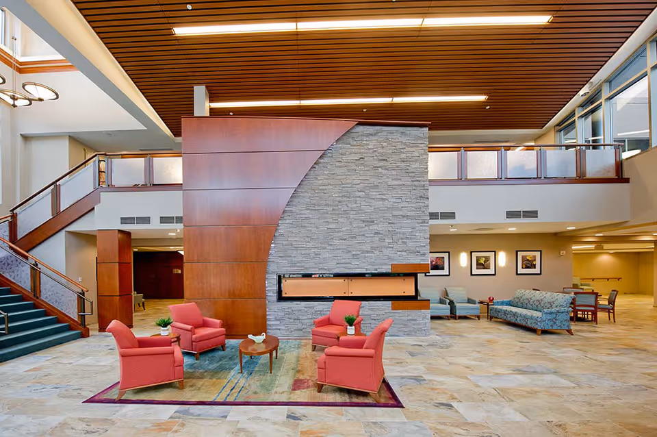 Spacious senior living facility common area with a modern stone and wood fireplace centerpiece. Four red armchairs are arranged around a small wooden coffee table on a colorful rug. To the right, there are additional seating options including a floral-patterned sofa and blue armchairs. A staircase with wooden railings leads to an upper level with glass panels. The ceiling features wooden slats and recessed lighting, and large windows allow natural light to fill the space.