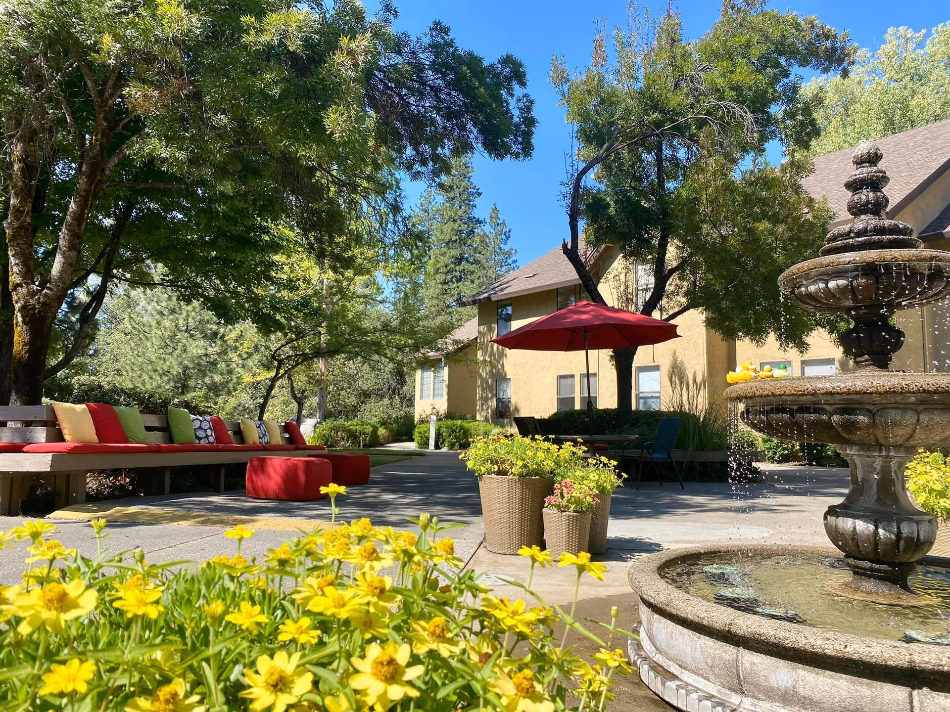 Sunny courtyard with a fountain, yellow flowers, a long bench with colorful cushions, and a red umbrella in front of a senior living building.