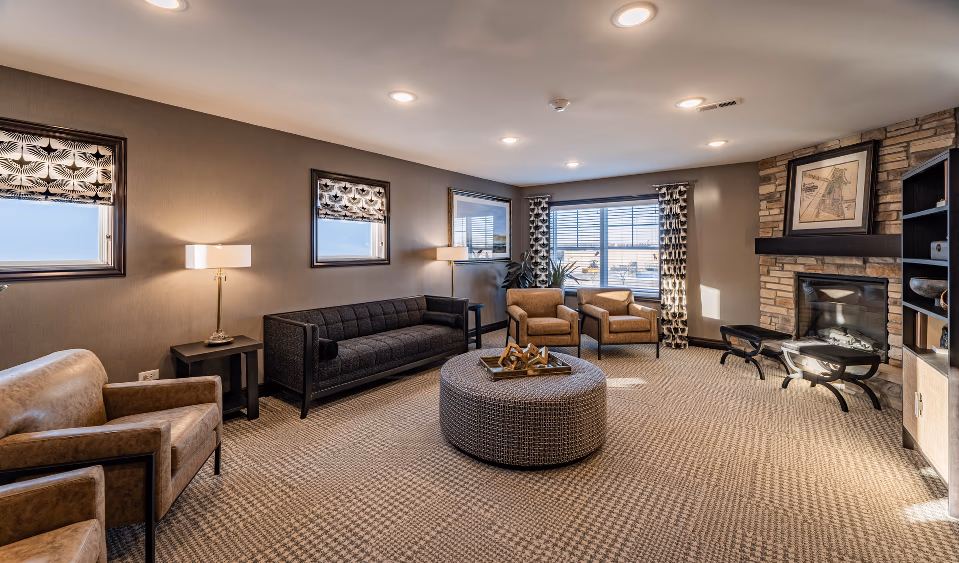 A spacious senior living common area with a patterned carpet, a dark gray sofa, two tan armchairs, a round ottoman in the center, and a stone fireplace with framed artwork above it. The room has multiple windows with patterned curtains and blinds, soft lighting from ceiling fixtures and lamps, and a built-in shelving unit on the right side.