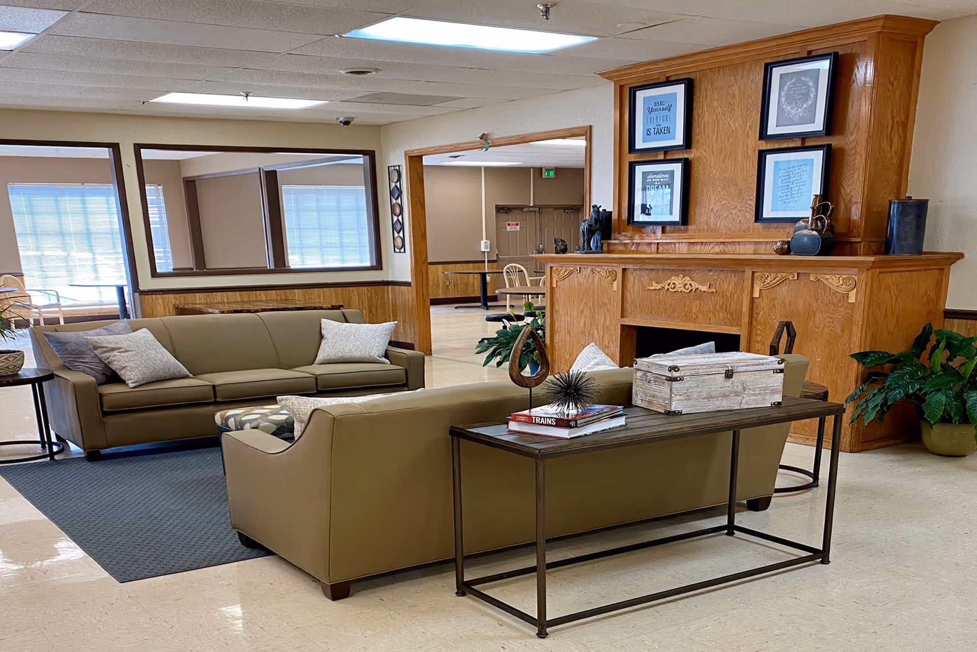 A cozy living room area in a senior living facility with two beige sofas arranged around a dark rug. A wooden console table behind one sofa holds decorative items including books and a small box. The room features a wooden fireplace mantel adorned with framed inspirational quotes and decorative objects. Large windows and open doorways provide natural light and views into adjacent rooms.