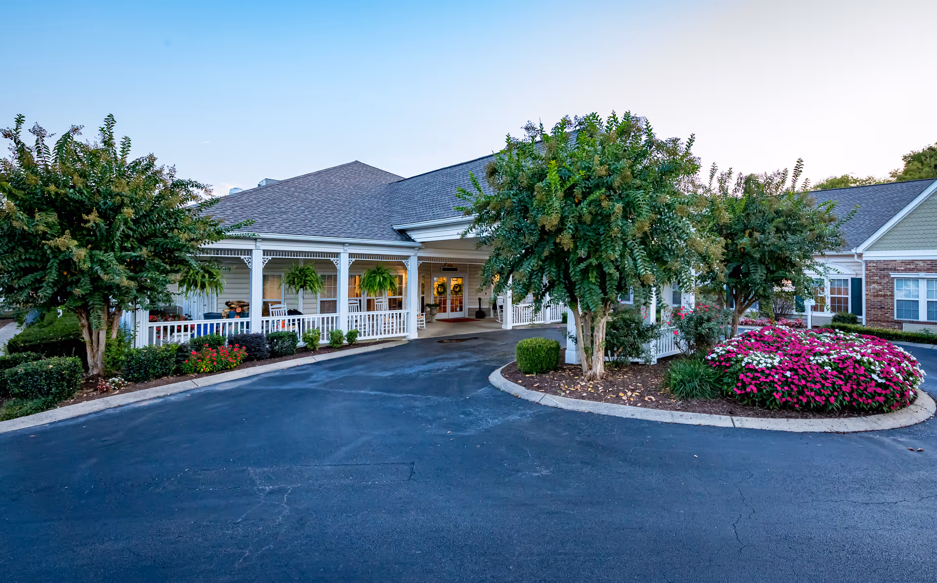 Exterior view of a senior living facility with a covered entrance, white railing porch, hanging plants, and landscaped flower beds with trees and colorful flowers.