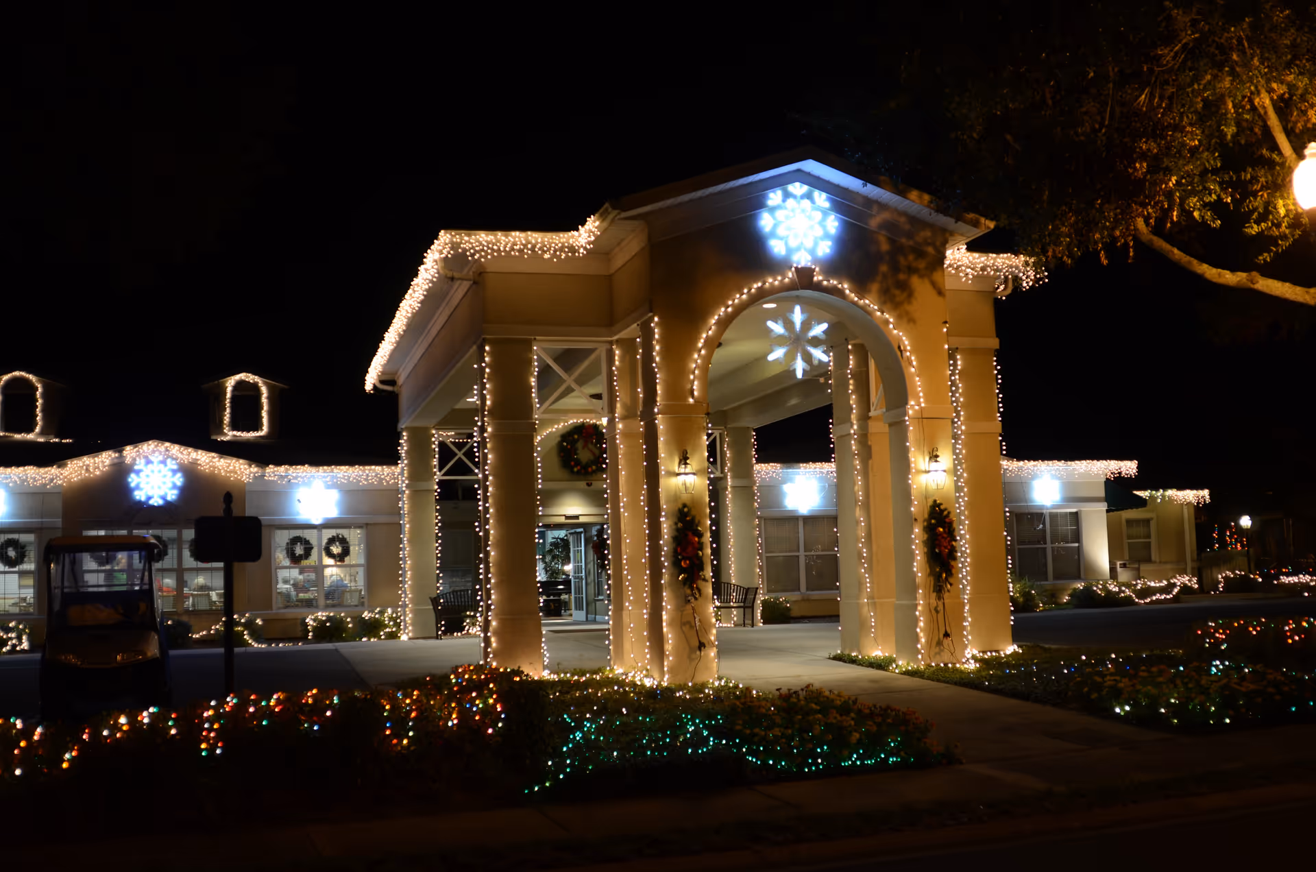 The entrance of The Oaks at Waterman Village Retirement Community decorated with white string lights and illuminated snowflake decorations at night. The building is adorned with holiday wreaths and festive lighting along the roofline and columns, with colorful lights in the landscaping.