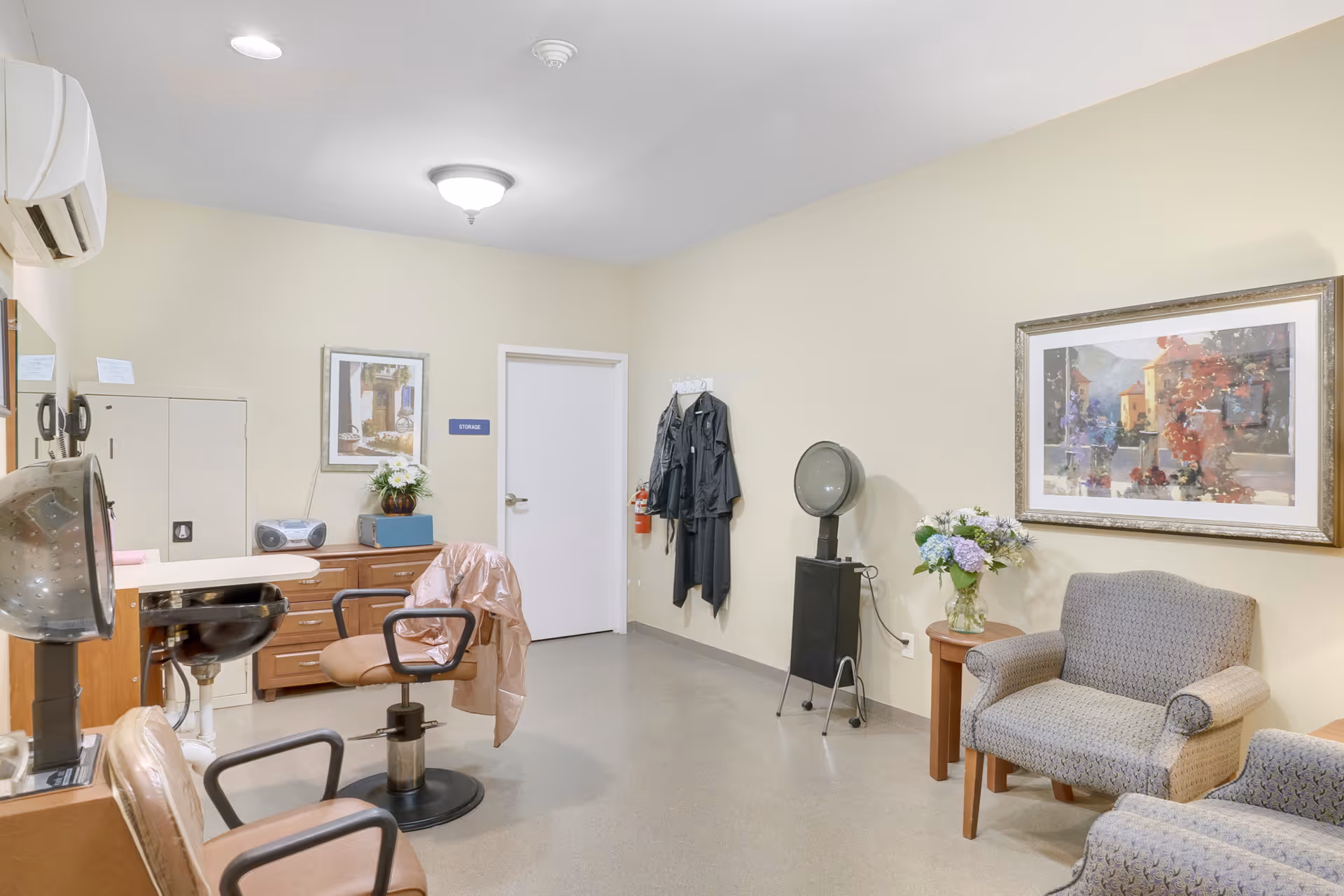 Interior of a hair salon room in a senior living facility with two salon chairs, a hair dryer, a small table with a vase of flowers, two armchairs, a painting on the wall, and a door labeled storage.