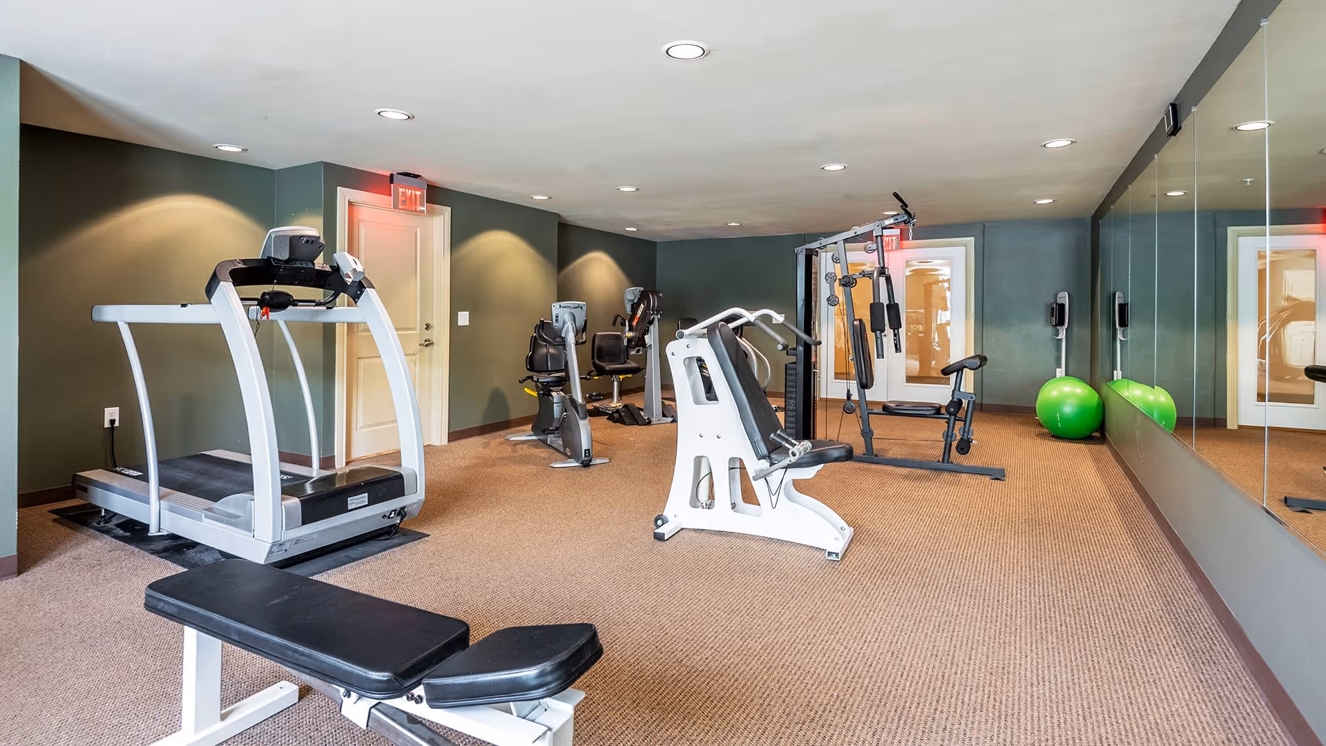 A fitness room with exercise equipment including a treadmill, stationary bikes, a weight machine, a bench, and two green exercise balls. The room has carpeted flooring, green walls, recessed ceiling lights, and a large mirror on one side.